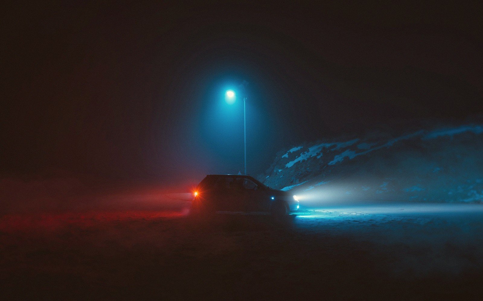 A car sits parked at night with red taillights glowing and headlights cutting through fog