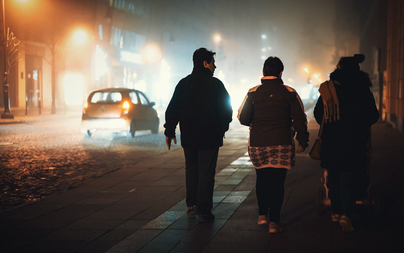 Pedestrians walk along a foggy, lamplit cobblestone street at night as a car approaches
